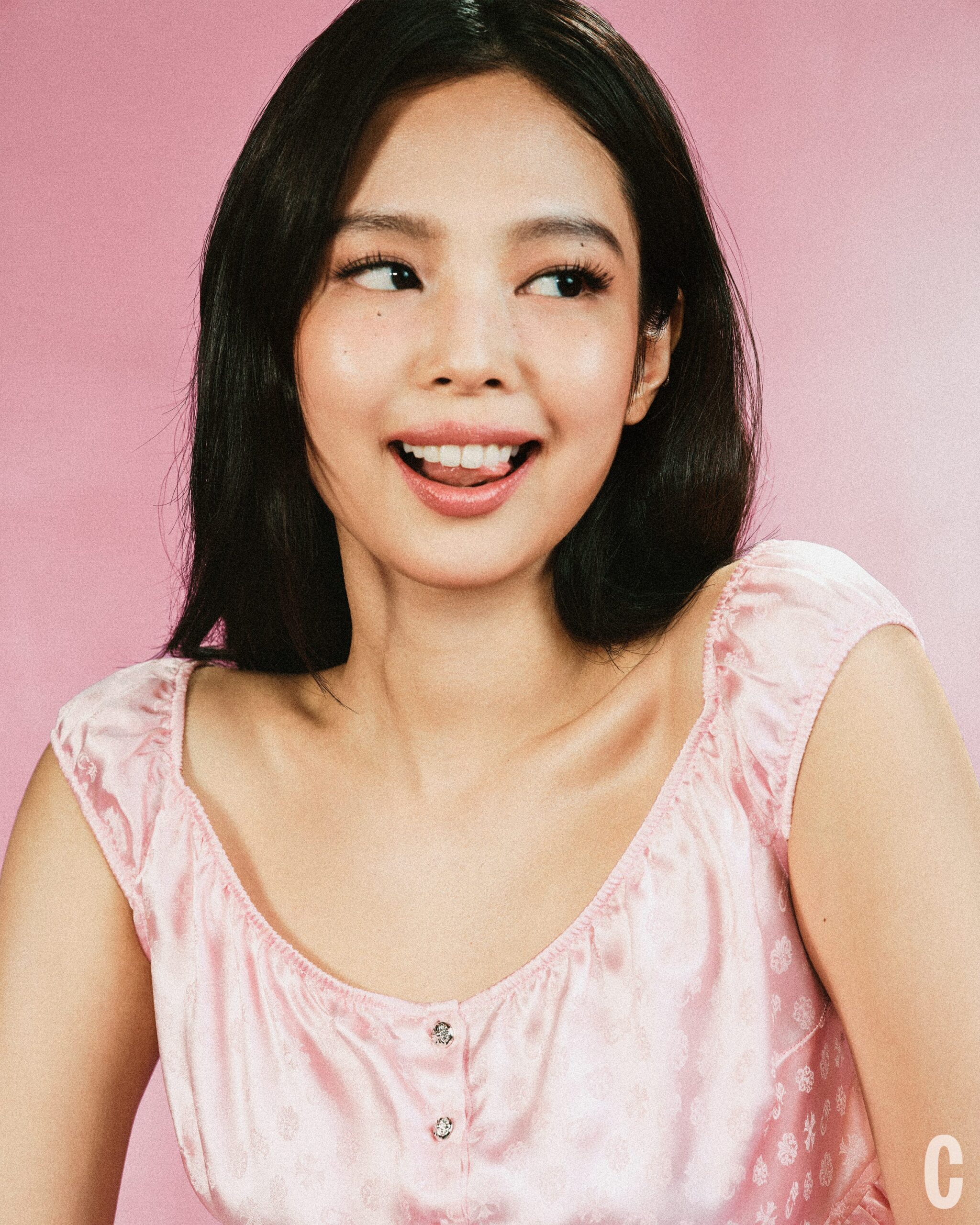 A young woman with long black hair, wearing a pink top, smiles playfully against a soft pink background.