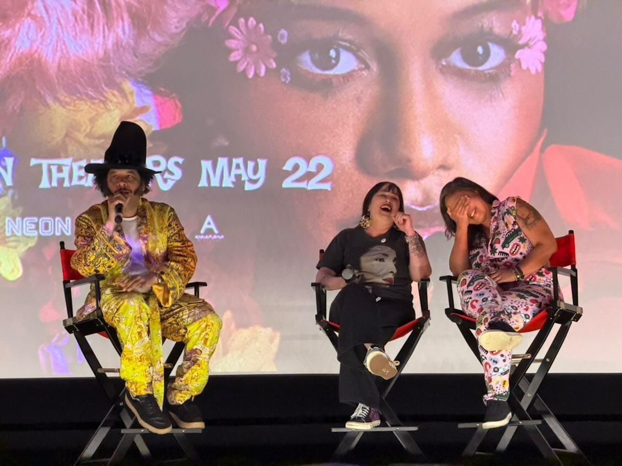 A Black man wearing a yellow outfit and very tall misshapen hat sits on a movie theater stage talking into a mic. To his left are two Asian women sitting in directors' chairs and laughing uproariously.