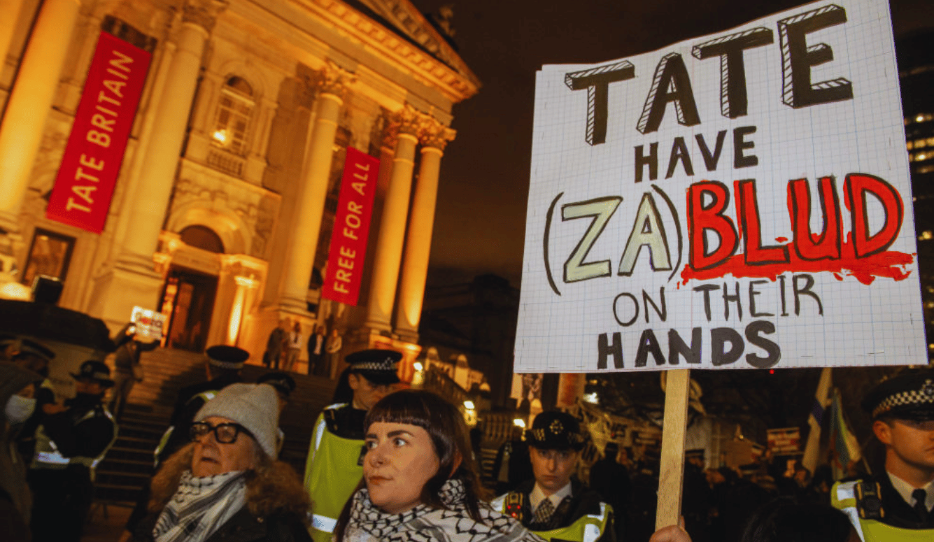 Protester holds a sign outside Tate Britain accusing the gallery of bloodshed as police and demonstrators stand nearby.