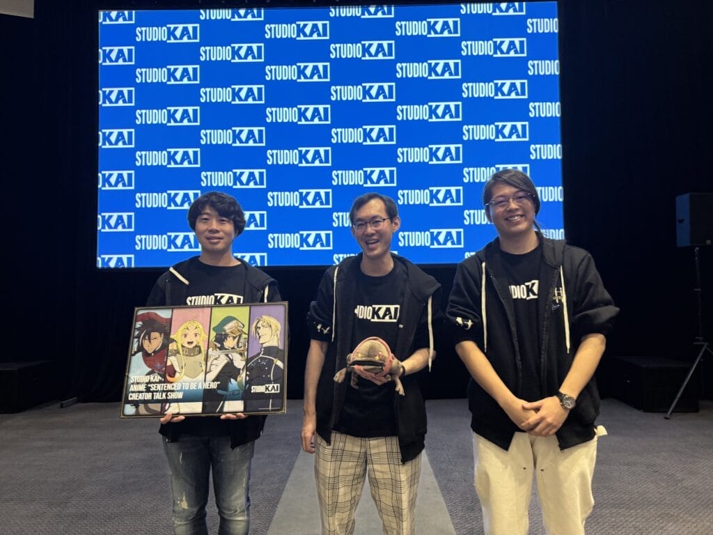 Photograph of the Sentenced to Be A Hero staff from Studio Kai, holding a visual for the anime.
