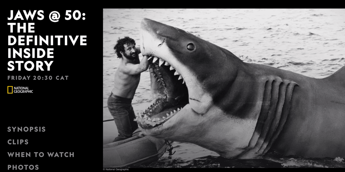 A man poses with his arm inside the open mouth of a giant mechanical shark in water. Text on the left reads: "JAWS @ 50: THE DEFINITIVE INSIDE STORY" celebrating the 50th Anniversary, with show details and National Geographic logo.