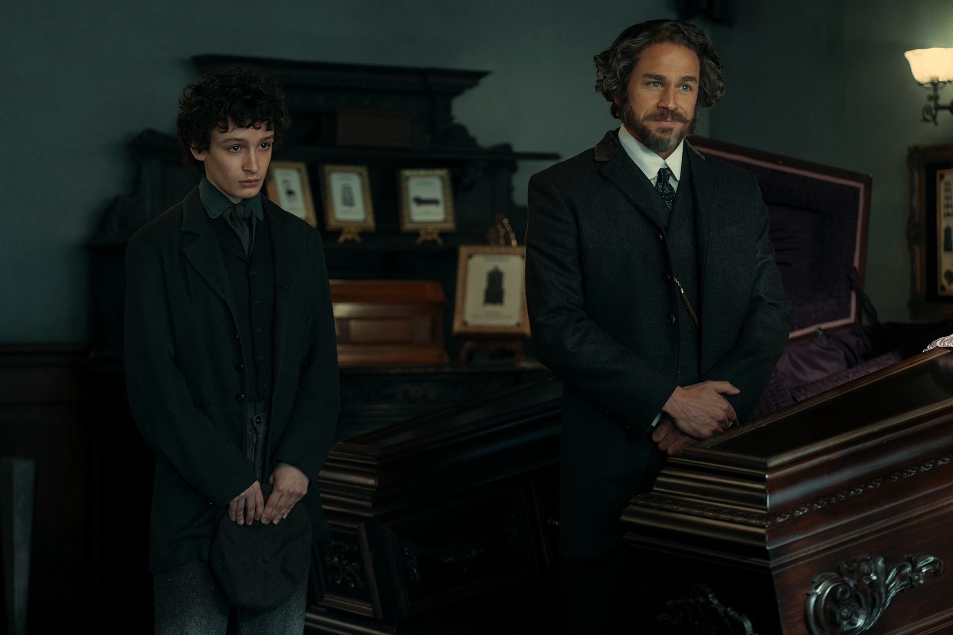 &Quot;Two Men In Dark, Old-Fashioned Suits Stand Beside A Polished Wooden Coffin In A Dimly Lit, Ornate Room Decorated With Framed Photos, Suggesting A 19Th-Century Funeral Or Wake Setting.&Quot;