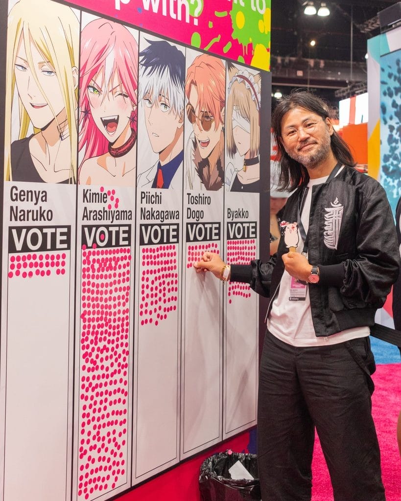 MARRIAGETOXIN director Motonobu Hori standing against a fan character voting display featuring the MARRIAGETOXIN cast.
