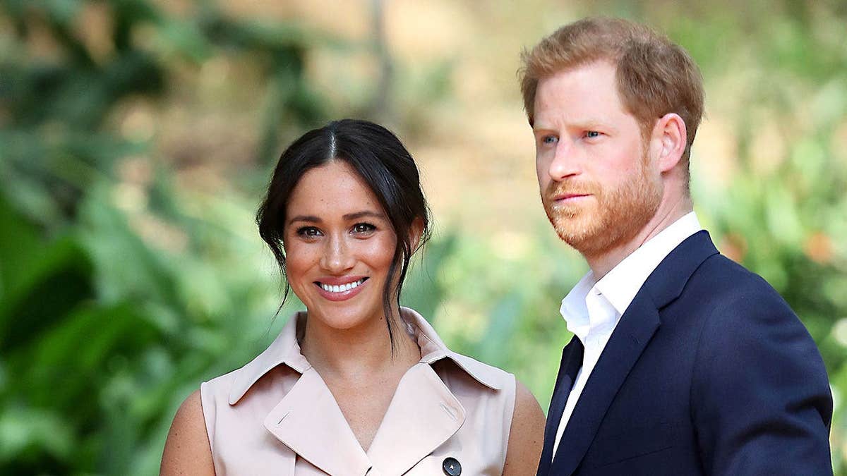 A close-up of a smiling Meghan Markle and a stern-looking Prince Harry against a forest backdrop.