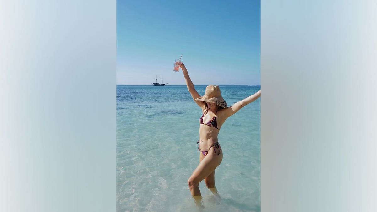 Michelle Monaghan raises both arms while standing in turquoise water wearing a colorful string bikini and wide-brimmed sun hat.