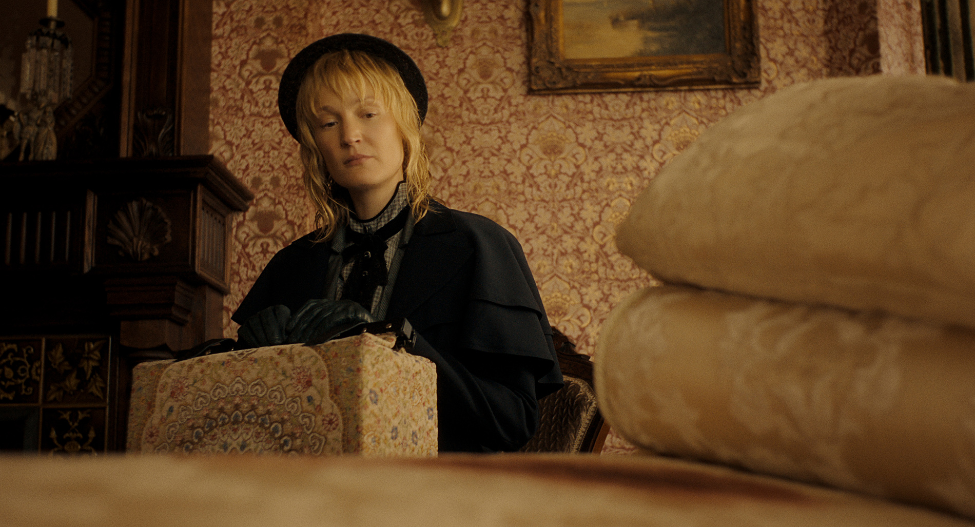 &Quot;A Woman In Vintage Clothing And Gloves Sits Indoors, Holding A Patterned Box. The Room Has Ornate Wallpaper, A Painting, And Folded Linens In The Foreground, Suggesting A Historical Or Period Setting.&Quot;