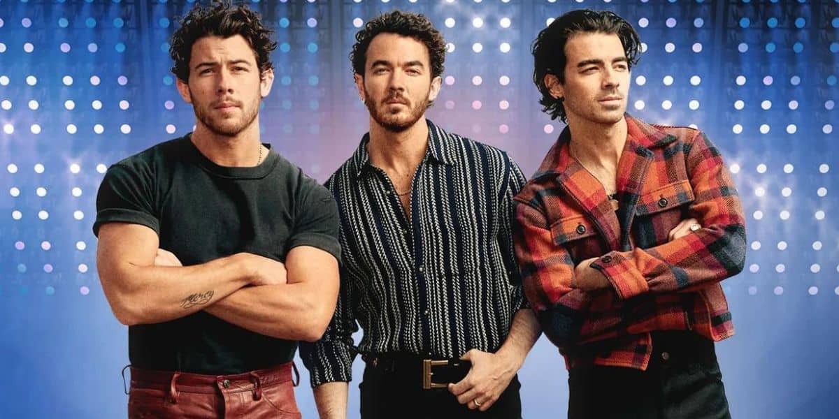 Three men stand side by side against a blue background with rows of white lights. Resembling the Jonas Brothers, the man on the left wears a black shirt, the center man a striped shirt, and the right man a red plaid jacket.