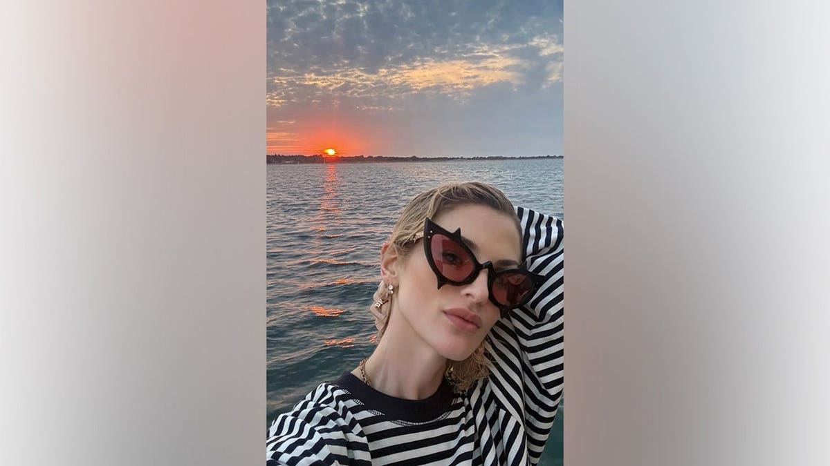 Kelly Osbourne takes a selfie in red-tinted cat-eye sunglasses and a black-and-white striped top with the sun setting over the water behind her.