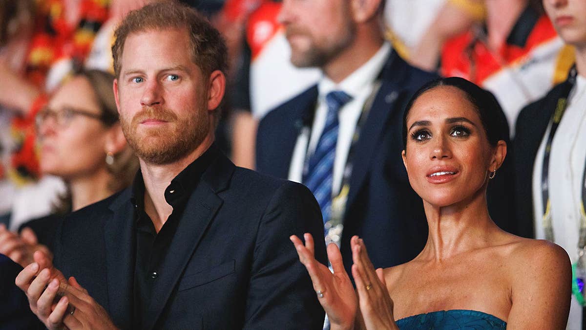 Prince Harry and Meghan Markle standing close to each other in an audience applauding and looking serious.