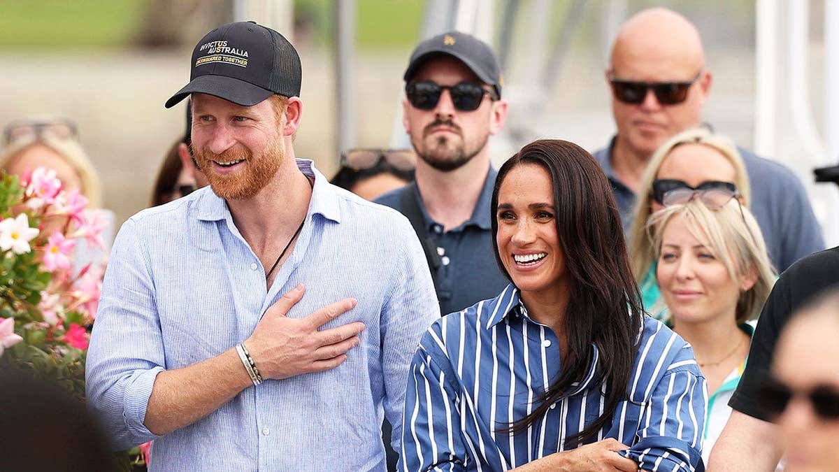 Prince Harry and Meghan Markle in casual clothes smiling and waving to a crowd in Australia.