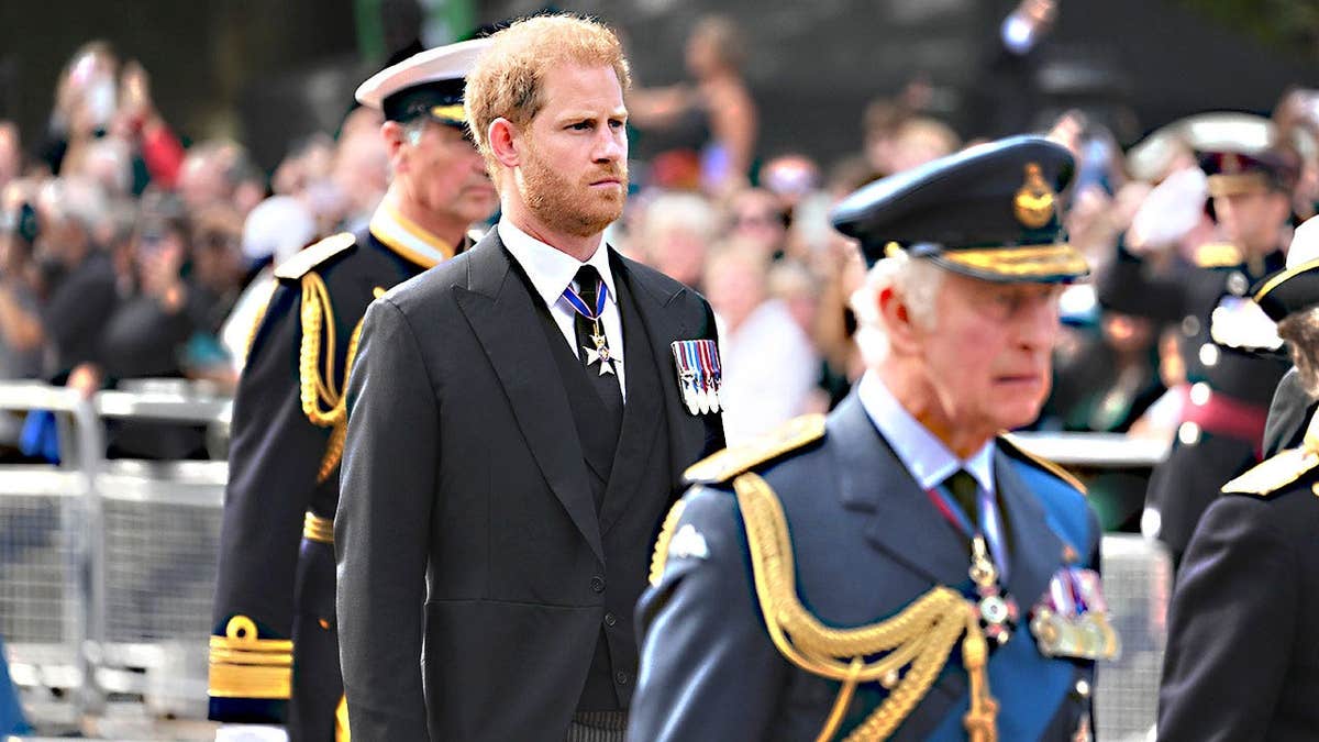 King Charles walking in a blue uniform looking somber in front of Prince Harry in a dark suit.