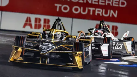 Getty Images Taylor Barnard drives in a black Formula E sports car with gold trim. There is another car chasing behind him which is black and white.