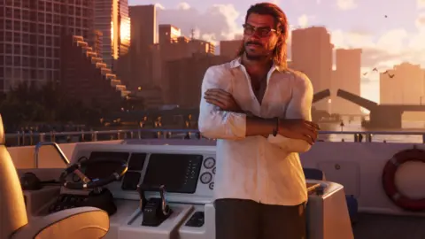 Rockstar A male character from GTA 6 in an open-collared shirt with slicked-back hair and sunglasses leans, arms folded, against the controls of a yacht as it coasts down a waterway flanked by skyscrapers. A road bridge opens in the distance as orange dawn light reflects of the glass fronts of the tall buildings. The computer-generated image is convincingly lifelike.