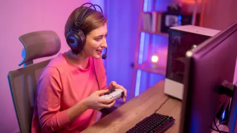 Getty Images A female streamer is sitting down at a desk looking at her computer, holding a white and black PlayStation 5 controller. She is wearing a pink jumper and has a black headset on.