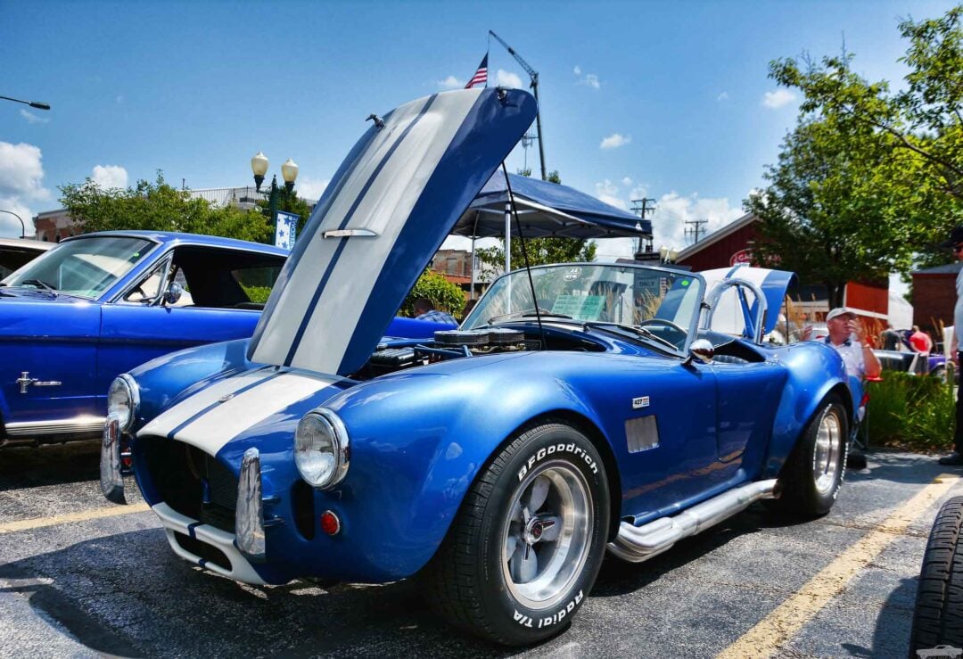 A blue classic car with white racing stripes is on display with its hood open at an outdoor car show. Another vintage car is visible in the background, and there are people and trees around under a sunny sky.