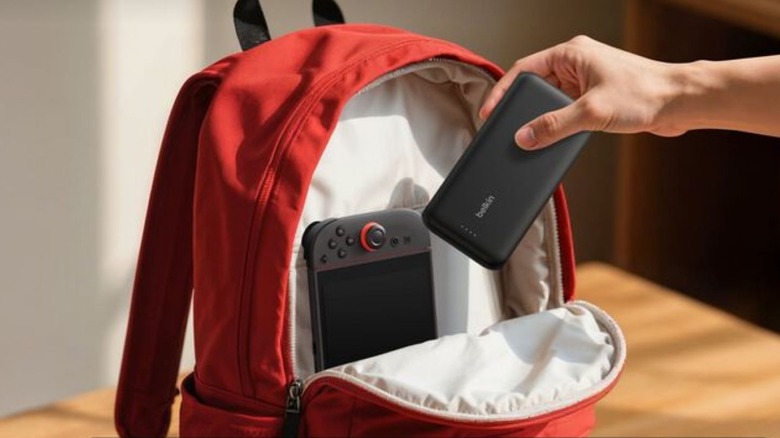 A hand putting a Belkin power bank into a red backpack next to a Nintendo Switch 2