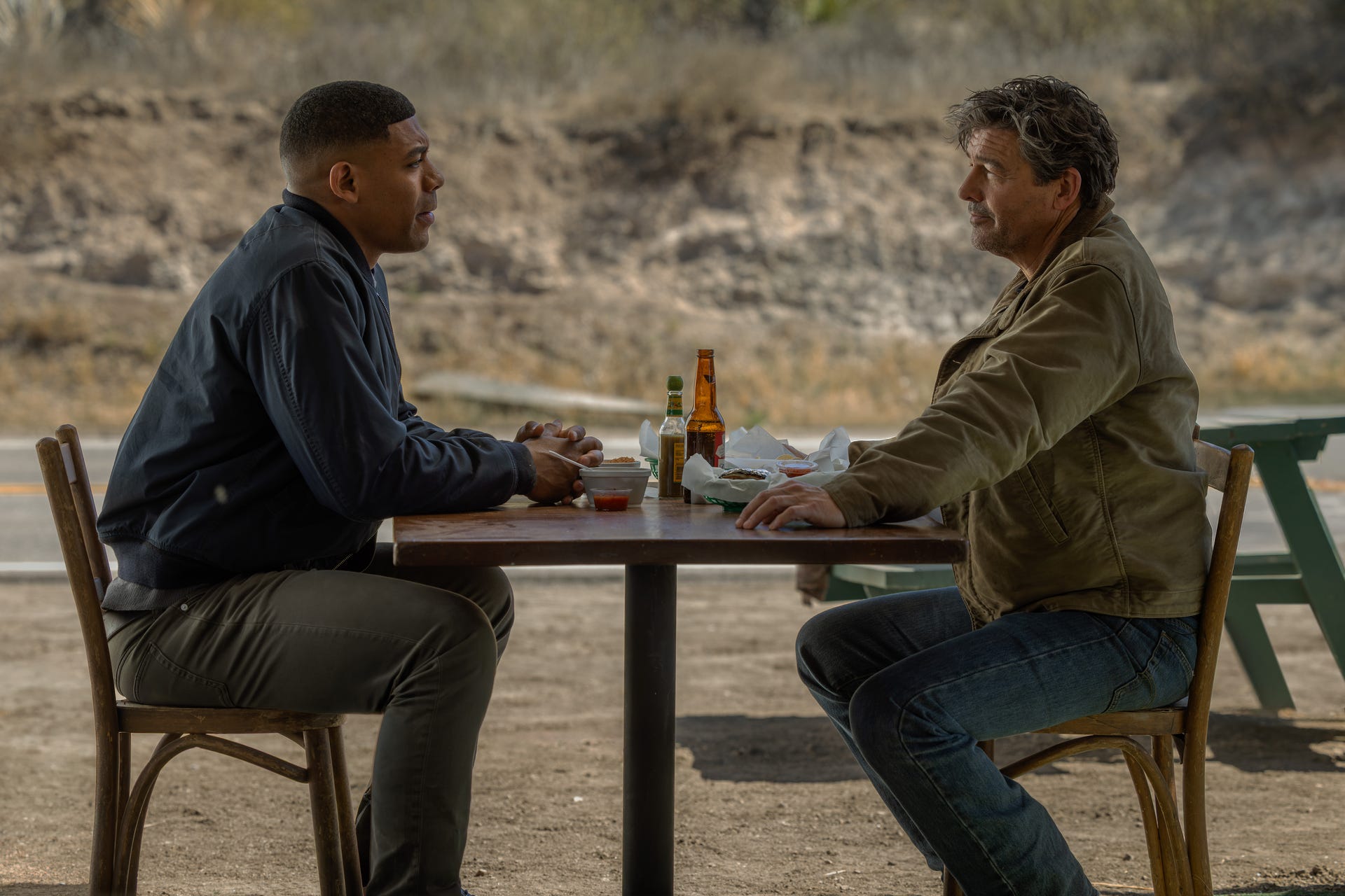 Two men engaged in a serious conversation over food and drinks at an outdoor table.