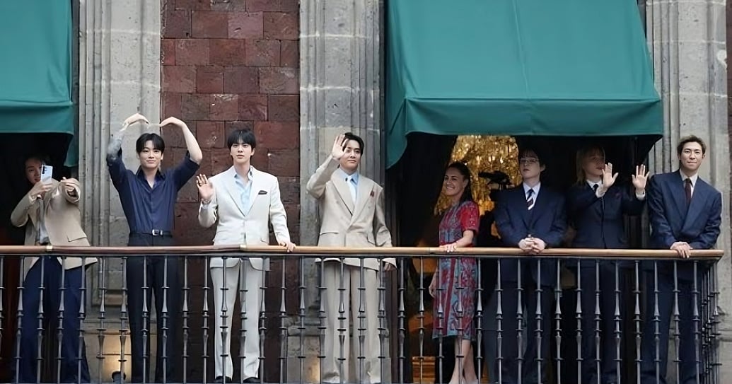 BTS members stand on a high balcony of a historic stone building. Jungkook makes a heart gesture over his head, Jin and J-Hope wave while wearing light suits, and Suga, Jimin, and RM stand on the right in dark suits waving to the crowd.