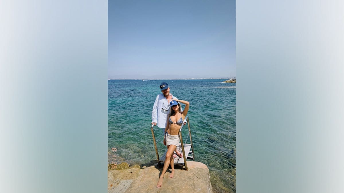 Kristin Cavallari wears a blue floral bikini top, a white crochet mini skirt and a navy baseball cap while posing beside turquoise Mediterranean water as Justin Anderson stands behind her in a white button-down shirt and a blue baseball cap.