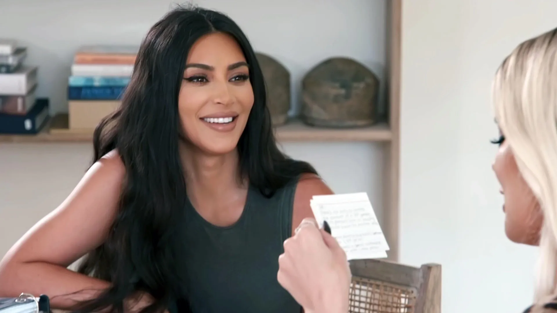 Kim Kardashian smiles while looking toward another woman holding a small white index card during a conversation.