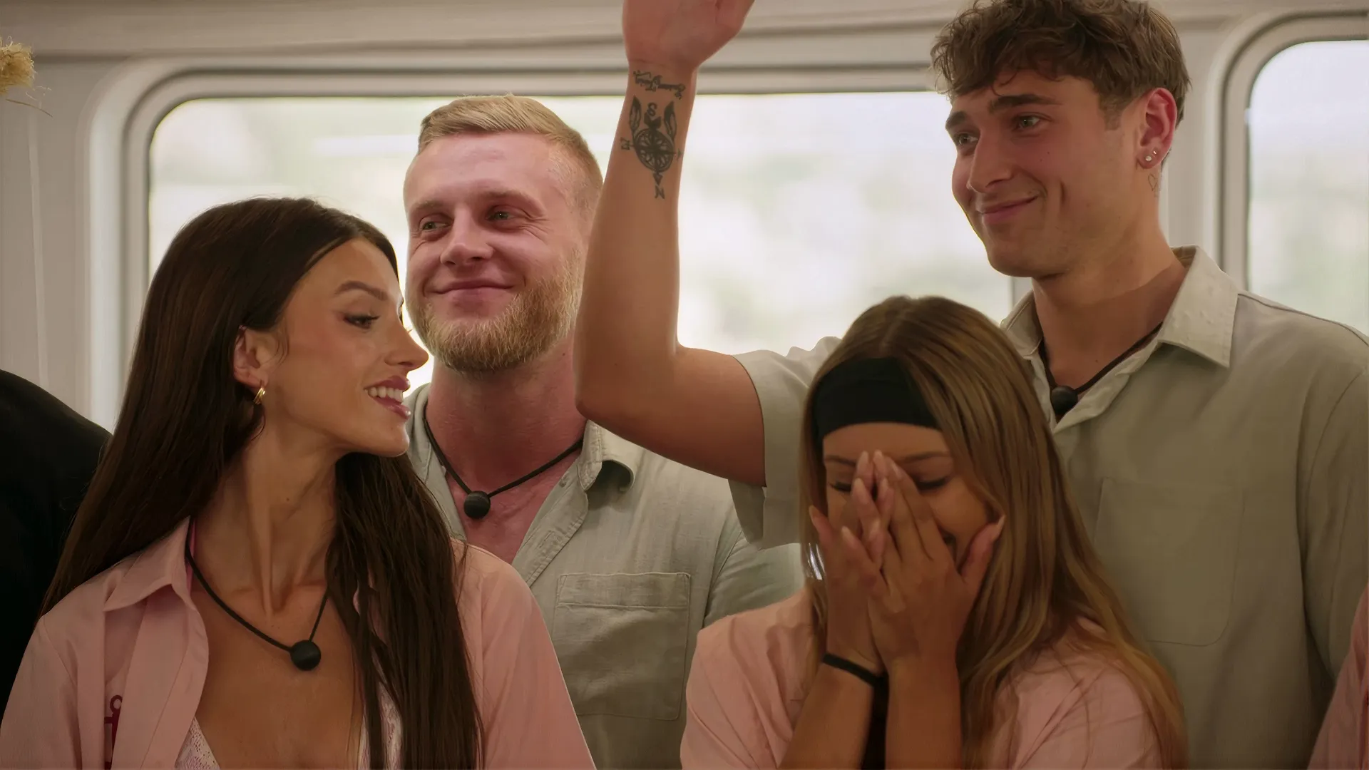 A group of four smiling young adults stands closely together, with one woman in the front right covering her face with her hands while a hand with a compass tattoo is raised above her.
