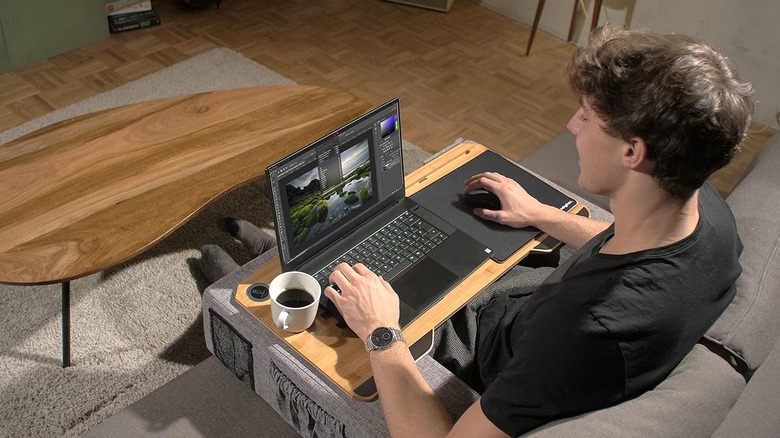 A man sitting on a couch using a laptop that's on a lap desk with a mouse and cup of coffee.
