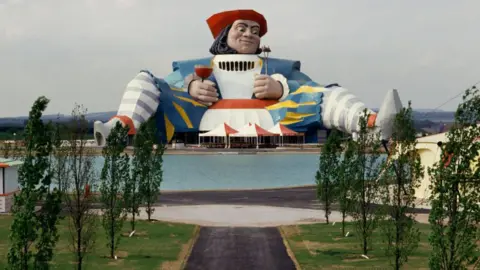 AFP via Getty Images A giant statue of a man, dressed in medieval clothing and folding a fork in one hand and a glass of red wine in the other, sits with his legs spread above a lake and trees in the former theme park
