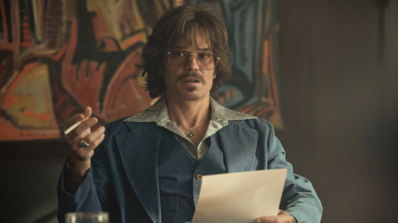 Timothy Olyphant with shaggy brown hair wearing a blue linen suit and waistcoat with a large open collared gray shirt underneath. He also has a mustache and has a cigarette in his right hand. He's holding a piece of paper in his left hand.