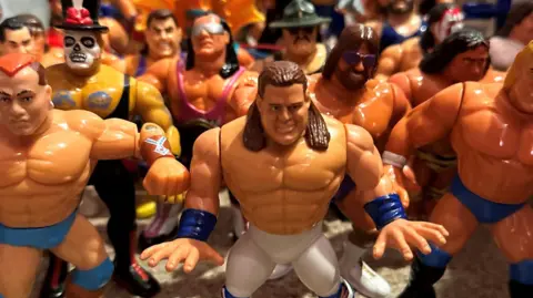BBC A collection figures of various wrestlers all standing in a huddle. They are bulky and muscly figures, rather cartoon like.