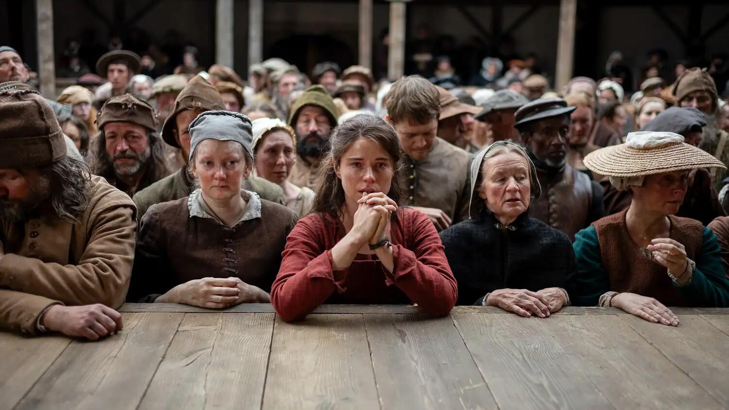 A woman in red stands at the edge of a stage surrounded by people dressed in period garb.
