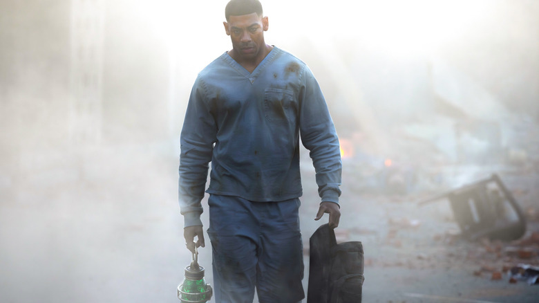 A Black man with a short haircut is wearing blue hospital scrubs. He's carrying a green lantern with a metal handle in his right hand and a black bag in his left hand. There is smoke and debris around him.