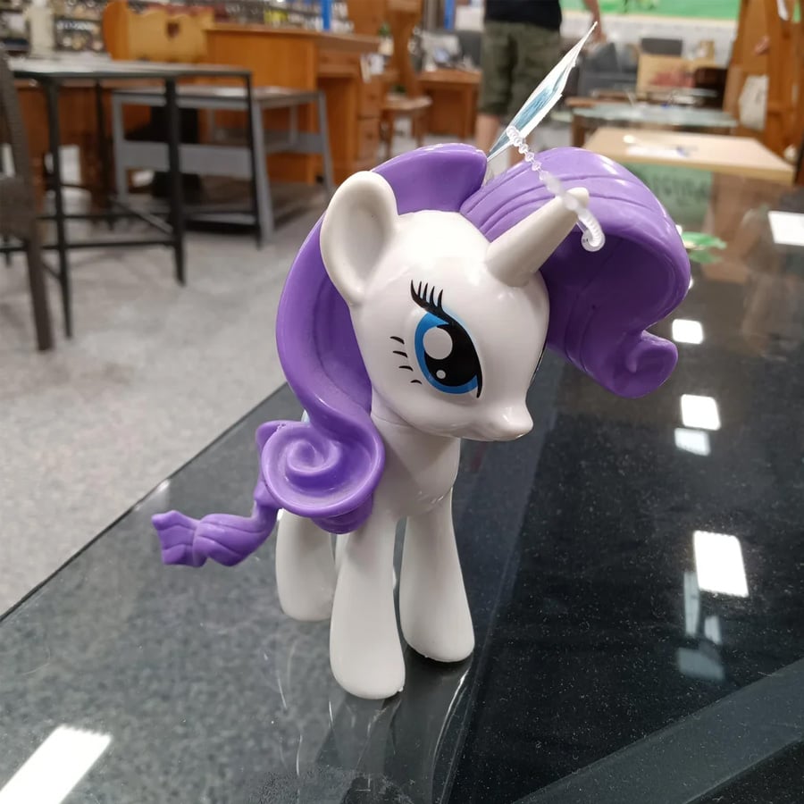 A toy unicorn pony with white body and purple mane stands on a glass surface in a store with wooden furniture in the background.