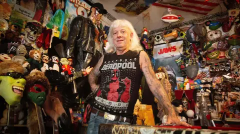 BBC Martin leans against a red pool table surrounded by a dense collection of pop-culture memorabilia. The room contains masks, stuffed toys, posters, model displays and a life-sized figure dressed in black standing behind them.