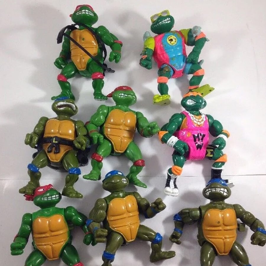 Eight Teenage Mutant Ninja Turtles action figures are arranged on a white surface. Two are dressed in colorful sports outfits while the other six wear standard gear with colored masks and belts.