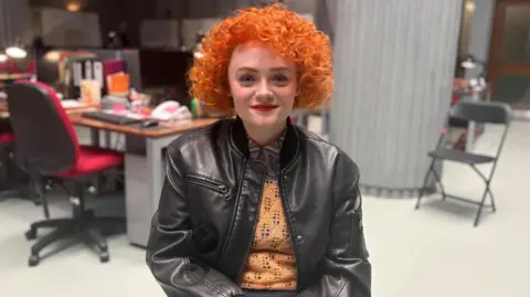 Leah Byrne, who plays DC Rose Dickson, sitting on a seat inside the building. A desk and chair can be seen over her right shoulder and a grey pillar is behind her left shoulder. She has short, bright orange curly hair and is wearing a black leather jacket over an orange top