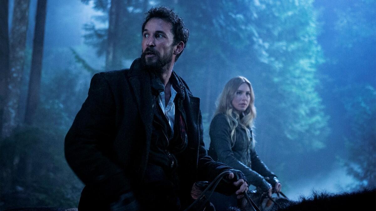 In a still from Falling Skies, a man with brown hair sits on a horse in a forest at night next to a blonde woman who is also on a horse.