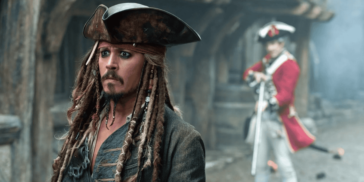 A man dressed as a pirate, with long braided hair, a tricorn hat, and a tattered coat, stands in the foreground looking to the side. In the blurred background of this 2025 Theme Park setting, a man in a red military uniform holds a musket and stands outside a wooden building reminiscent of Pirates of the Caribbean.