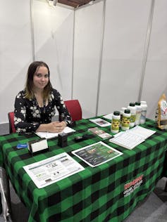 A woman sits behind an information booth with a checkered green and black tablecloth and some information and products on top.