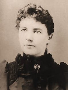 Black and white portait of a woman with her hair in a bun wearing a 19th century-style dress with a high ruffled neck.