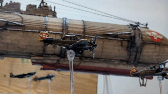a custom game of Gaslands themed around Crimson Skies - a WW2 era fighter aircraft flies past a massive blimp