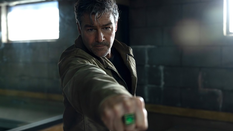 A middle-aged man with graying hair points his fist at something off-camera, a green ring is on his middle finger. He's wearing a brown jacket, and is stood in what looks like a basement.