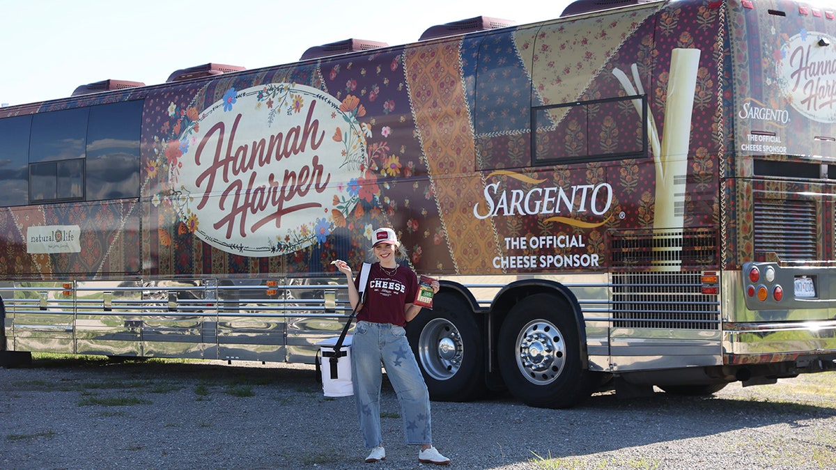 Hannah Harper poses in front of a Sargento-branded tour bus while holding cheese snacks outdoors.