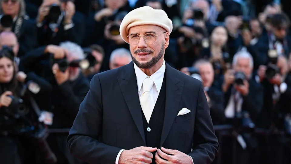 John Travolta in a white beret and glasses, wearing a black suit and white tie, poses on a red carpet with photographers in the background.