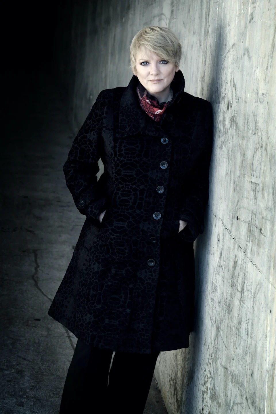 Alison Arngrim in a black coat leans against a concrete wall in a dramatically lit portrait