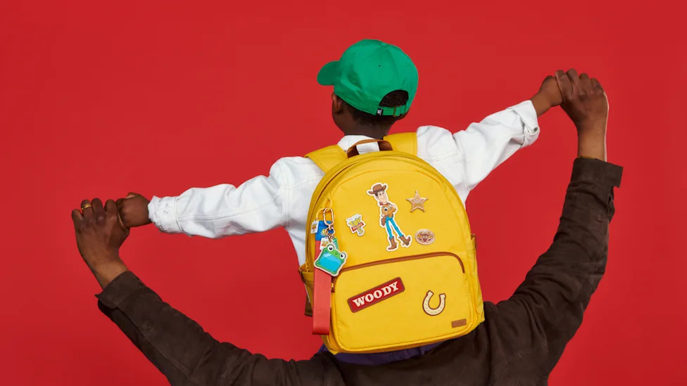 Person wearing a yellow Away Toy Story 5 Woody backpack covered in character patches against a red background