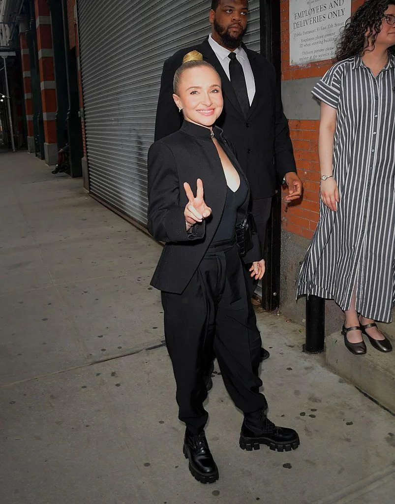 Hayden Panettiere, seen on May 19, smiling and posing while doing a peace sign with her fingers on a New York City street. Her blonde hair is tied in a bun and she wears a black blazer, with a black top, black trousers and black boots.