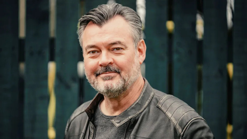 Grant Stott, with a grey beard and wearing a leather jacket, standing against a garden fence