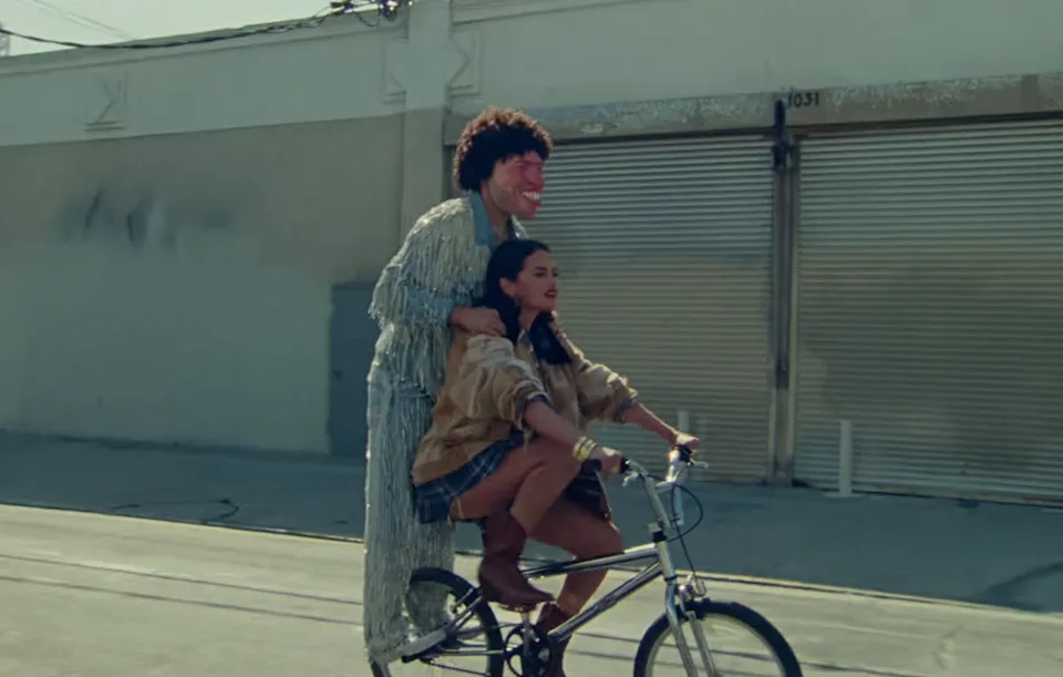Benny wearing a large, exaggerated mask and sparkly outfit, riding a bike with Selena in a music video