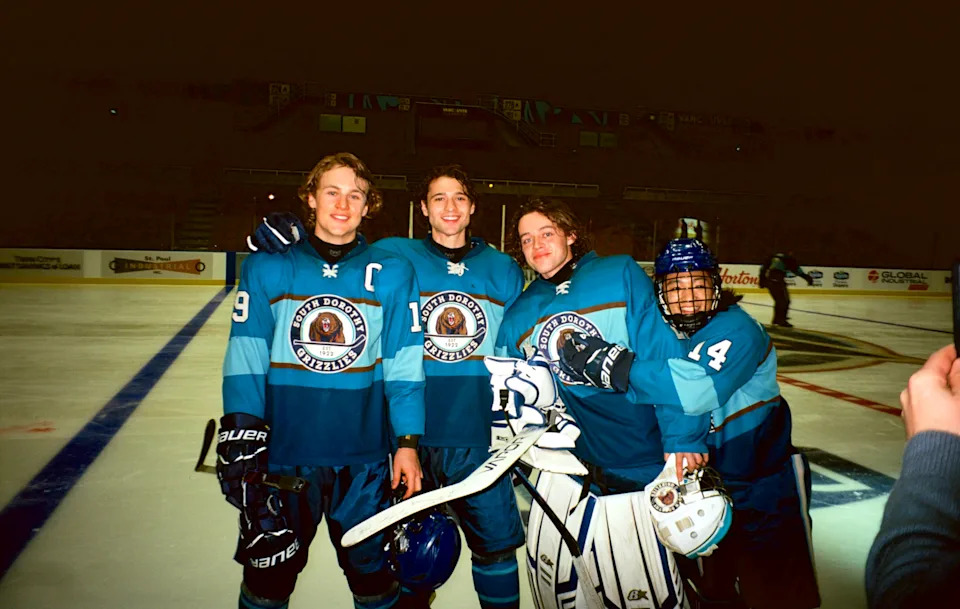 Four hockey players in blue team uniforms stand on an ice rink, smiling and posing for a group photo