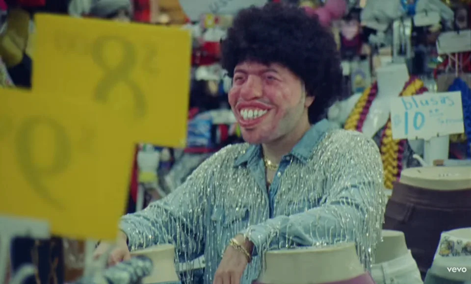 Benny wearing a large, exaggerated mask and sparkly outfit in a music video
